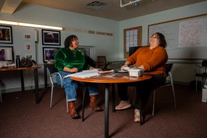 Kimeco Roberson (left) and Meida McNeal (right) laughing as they sit in their office at the First Church of the Brethren reviewing Honey Pot Performance archive materials. Kimeco is wearing a green velvet top, blue jeans, and brown boots. Meida McNeal is wearing a dark orange sweater top, dark brown pants, and pink sneakers. Image by Tonal Mondae.
