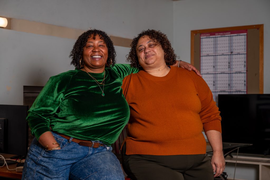 Kimeco Roberson (left) and Meida McNeal (right) lean against an office desk and smile at the camera. Kimeco’s left arm is around Meida as her hand sits on Meida’s left shoulder. In the background is a partial view of a large wall calendar and tv. Image by Tonal Simmons.