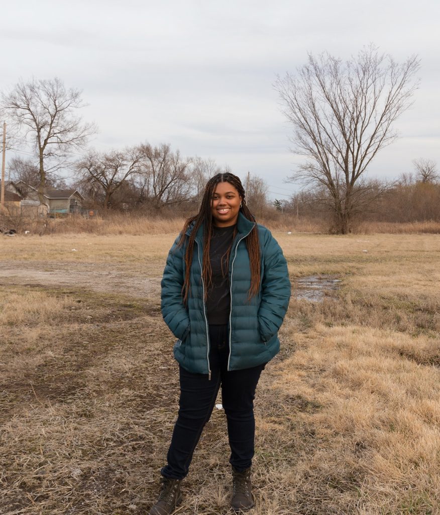 A photograph of Allena Marie Brazier. She is standing in the center of field wearing a turquoise wither coat with a black shirt underneath with black pants and boots. Image courtesy of the artist.