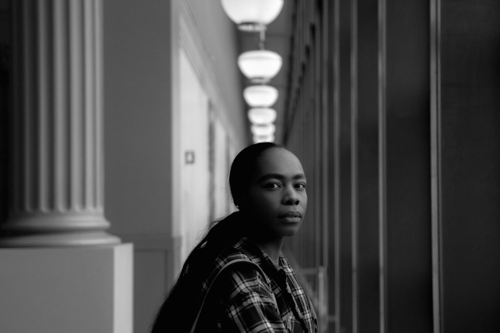 Image: Black-and-white portrait of bria royal in semi-profile, flanked on either side by columns and beams. Above her head are a series of globular light fixtures. Photo by Sebastián Hidalgo.