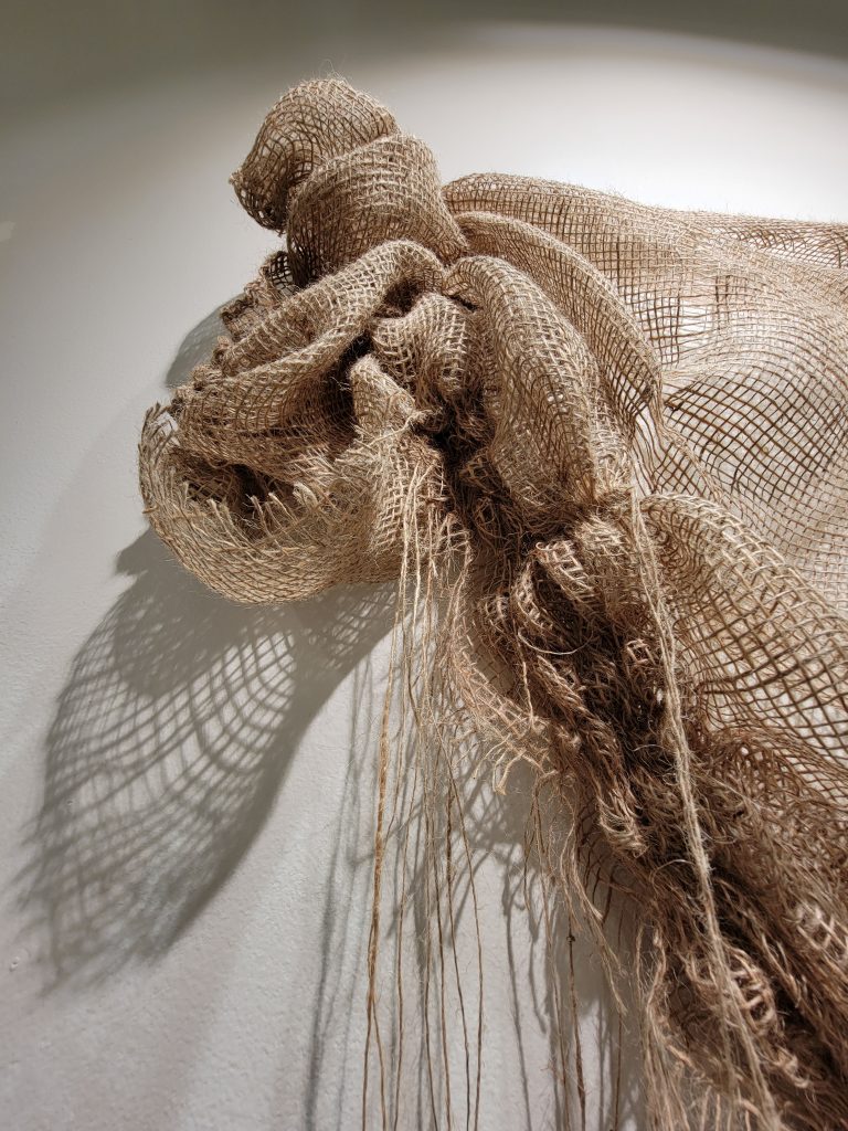 No Strings Attached art series by by Allena Marie Brazier, burlap, 2023. Detail of No Strings Attached. Shadows of burlap are cast onto the wall. Unravelled pieces hang. Image courtesy of the artist.