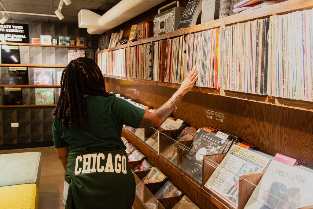 Dr. ShaDawn Battle chooses records inside the National Public Housing Museum. The walls are lined with warm-toned wood shelves and covered in records. Dr. Battle has a dark green sweatshirt wrapped around her waist with the word CHICAGO in bright white letters. Image by Joshua Clay Johnson.