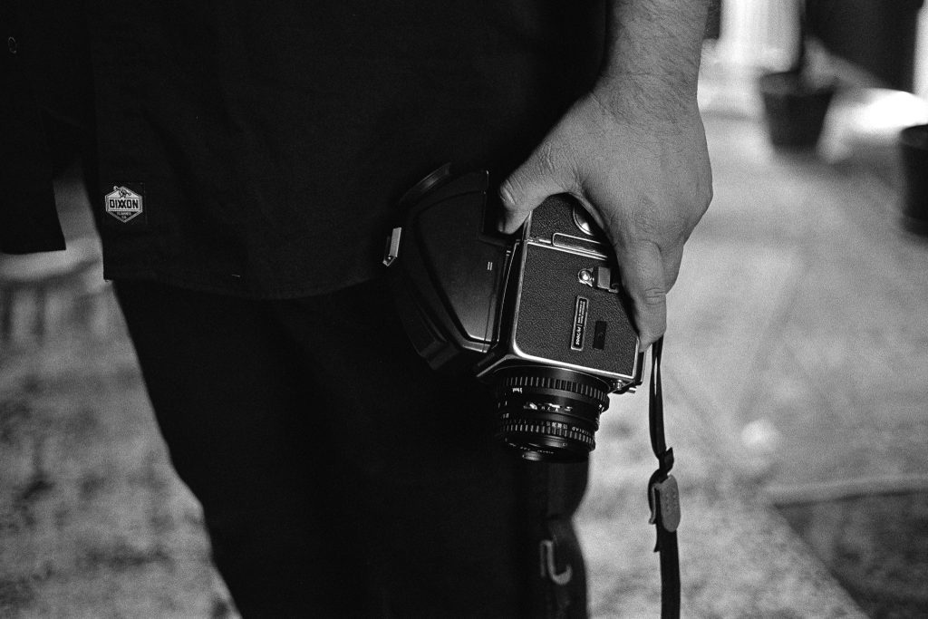 A black and white capture of photographer Carlos Martinez holding his vintage camera in his hand down by his hip. Photo by Genesis Falls.