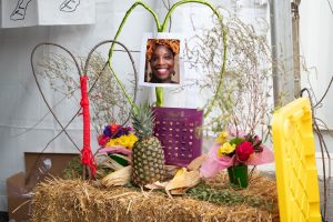 Image: An altar made of hay honors Patrisse Cullors, artist, activist, and co-founder of Black Lives Matter. She was unable to attend The Circus after her home was unlawfully raided by police. Created by her co-creators from the Spell Casting booth, the altar features two small, colorful flower bouquets in green vases placed at each end. Between them sit a pineapple, an ear of corn with its husk, and a purple box of Lindt chocolates. Two hearts made from sticks rest nearby—one wrapped in green yarn and the other in red. A printed photo of Patrisse Cullors is placed at the center of the green heart. Photo by Tonal Mondae.