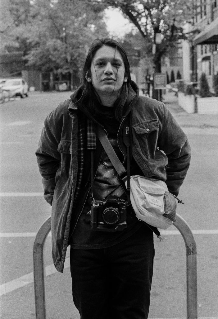 A black and white film capture of photographer Josue Cuyun standing in the middle of the frame with his camera hanging on his neck. Photo by Genesis Falls.