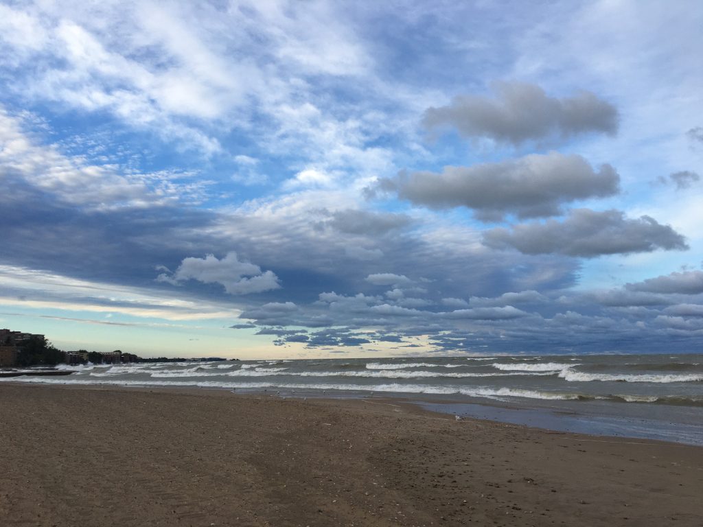 A landscape photo of Loyola Beach looking out at the white-capped waves of Lake Michigan as they come to shore. The light blue sky is covered in wispy, white, feathery clouds high above while lower are more chunky, fluffy, grey ones. Photo courtesy of Emily McClanathan.