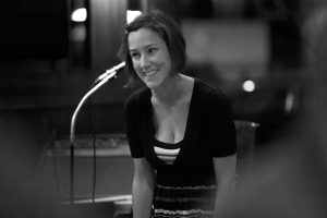 Image: Maggie Andersen who has short, dark hair, wears a striped tank top under a black v-neck is shown smiling while seated on a wood chair during a conversation at one of her first GiftLabs events. Courtesy of Maggie Andersen.