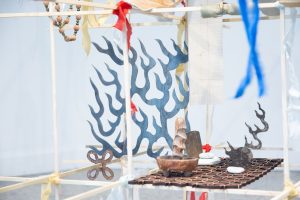 A wooden structure fabricated from rods is adorned with strips of blue and red ribbon, beads, metallic cutouts that resembles flames, stones, a ceramic object, and an ornamental metal panel.