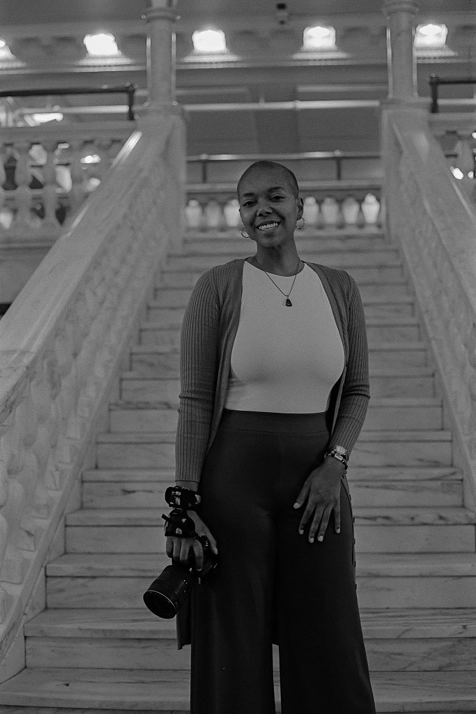 Black and white captures of photographer Sam Wilson holding her camera in her hands while smiling and standing at the bottom of a white staircase. Photo by Genesis Falls.