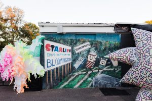 Image: Two big colorful balloons in the shape of stars decorate the entrance of Star Lite Drive-In Theatre along with a painted mural of animated consession stand treats dancing towards a screen. Photo by Garrett Ann Walters.
