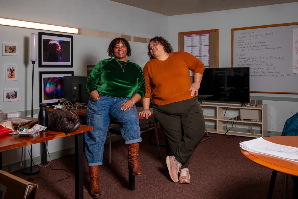 Kimeco Roberson (left) sits partially on an office desk with her left leg while standing on the right. Meida McNeal (right) is leaning on the table with her right hand while looking directly at Kimeco. Behind them, to the left, are framed photos from performances and events hanging. To the right is a white board with a to-do list, wall calendar, and tv with stand. Image by Tonal Simmons.