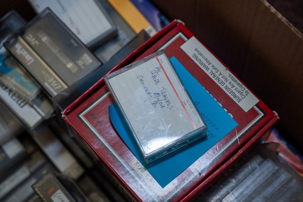 A small cassette tape sits on top of a red cigar box and a blue sticky note. Written on it is: Race Travels - 6/11/04 - Camera 2. In the background are other archived cassette tapes. Image by Tonal Simmons.