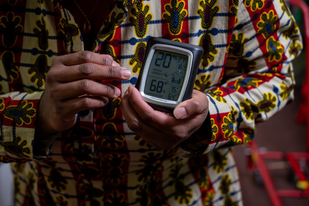 A close-up shot of Siobhan holding a black and gray digital hygro thermomerter in the archival room. It shows the temperature of 68 degrees farenheit and humidity of 20%. These are part of Siobhan’s practice to help Honey Pot keep track of warmth and moisture as they continue to organize their archive. Image by Tonal Simmons.
