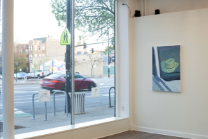 Image Caption: An installation view from inside the gallery, looking out through a large glass window onto an urban street scene. Two drawings are hung on the lower portion of the window, and one vertical painting on the right side wall depicts an el sombrero left inside a car, seen through the front windshield. Image courtesy of the artist.